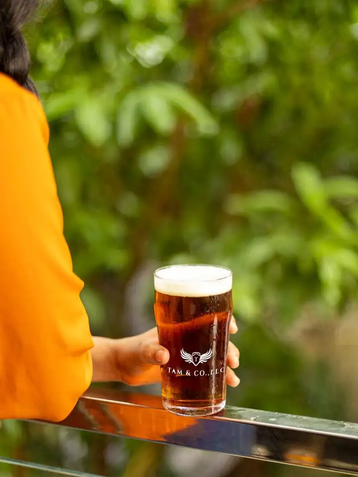 A glass of Tam & Co. beer held up outdoors against green leaves